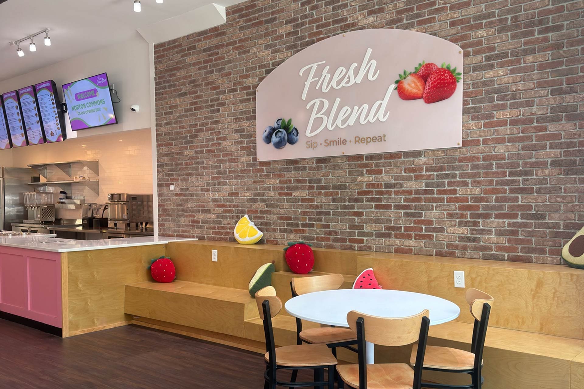 Cafe interior with a brick wall and a large sign reading 'Fresh Blend' featuring strawberries and blueberries; cozy seating with fruit-themed cushions.